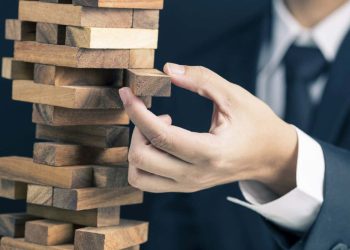 man in business suit pulls a piece out of wobbly wooden tower