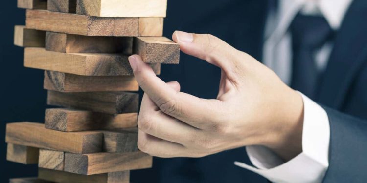 man in business suit pulls a piece out of wobbly wooden tower