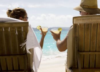 people sit in two wooden beach chairs facing the Caribbean ocean holding drinks and making a toast