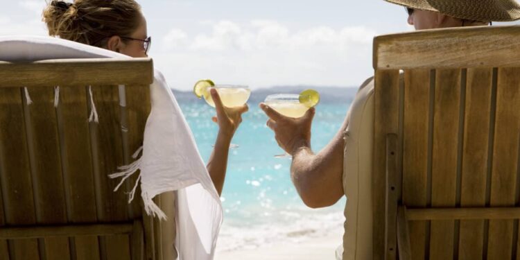 people sit in two wooden beach chairs facing the Caribbean ocean holding drinks and making a toast