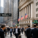 Crowds walk through Wall Street beside the New York Stock Exchange and its American flags.